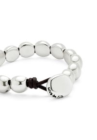 Pulsera de cuentas de metal en tono plateado con formas pulidas y redondeadas. Presenta un cordón de cuero oscuro y una etiqueta de firma en una de las cuentas.