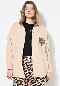 Camicia texturizzata color beige chiaro con maniche lunghe, tasche frontali e una grafica di una tigre nera sulla sinistra. Abbinata a pantaloni con stampa leopardata in crema e nero.