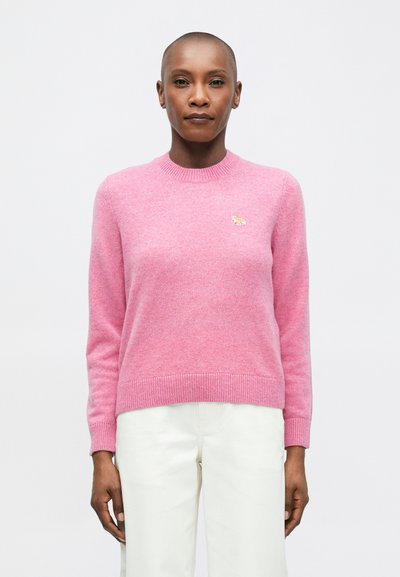 Une personne aux cheveux courts porte un pull long rose avec un petit animal brodé sur la poitrine et un pantalon blanc, se tenant devant un arrière-plan blanc.