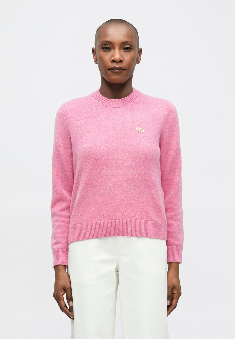 Osoba z krótkimi włosami nosi różowy sweter z długim rękawem z małym haftowanym zwierzątkiem na piersi oraz białe spodnie, stojąc na tle białym.