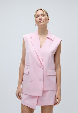 Donna con capelli biondi che indossa un blazer doppiopetto rosa senza maniche e una gonna asimmetrica abbinata, in piedi contro uno sfondo chiaro uniforme.