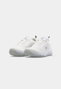 Chaussures de sport blanches avec une surface texturée, un design à lacets, des semelles gris clair, et un logo sur la languette. Équipées d'un onglet au talon.