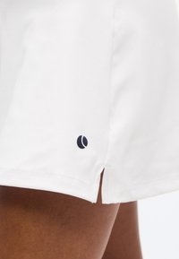 Κοντινό πλάνο λευκής φούστας με μικρή πλευρική σχισμή και μαύρο κυκλικό λογότυπο κοντά στο τελείωμα πάνω σε καφέ δέρμα.