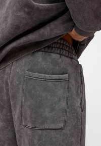 Grijze, gestructureerde joggers met een grote zak en een elastische tailleband, zijaanzicht met een losvallend bovenstuk dat gedeeltelijk zichtbaar is.