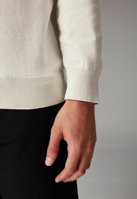 Sudadera de color beige claro con puños acanalados, diseño ajustado, mostrando una mano contra un fondo oscuro, destacando la textura y los detalles de la tela.