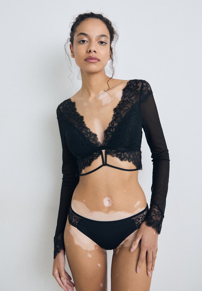 Juodas nėriniuotas liemenėlis su giliu V formos iškirpimu ir ilgomis permatomomis rankovėmis, derinamas su atitinkamais juodais kelnaitėmis su nėriniais. Lygus tekstūros paviršius ir subtilūs detalės.