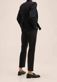 Veste de costume ajustée noire et pantalon fuselé, en tissu lisse, associés à des mocassins noirs. Vue de dos, détails minimalistes.