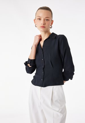 Donna con i capelli raccolti, che indossa orecchini a cerchio d'argento, una camicia nera con bottoni e maniche arrotolate, e pantaloni bianchi plissettati su sfondo bianco.