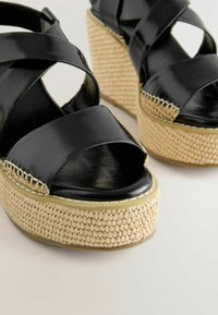 Sandales compensées en cuir noir avec larges brides croisées et semelles plateforme tissées beiges sur fond blanc.