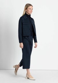 Marineblauwe cropped jas met twee voorzakken, gecombineerd met bijpassende wijde broek. Model draagt beige blokhak schoenen. Eenvoudig, strak ontwerp.