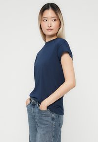 Junge Frau mit langen glatten Haaren, die ein navyblaues T-Shirt und blaue Jeans trägt, steht mit den Händen in den Taschen und schaut zur Seite.