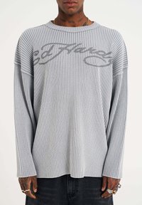 Light gray ribbed long-sleeve shirt with a crew neckline and dropped shoulders. Features contrasting dark gray text logo across the chest.