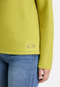Zielona sweter z prążkowanej dzianiny w odcieniu limonki, z zaokrąglonym dołem, z błyszczącym logo marki na przodzie i długimi rękawami.