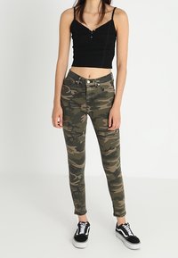 Kamouflage-mönstrade skinny jeans i nyanser av grönt och brunt, med medelhög midja och åtsittande silhuett, ihop med en svart crop top och sneakers.