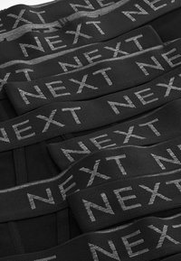 Benzi elastici negri cu o finisare texturată, având un logo argintiu repetitiv "NEXT". Aranjate într-un model stratificat.