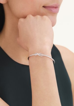 Bracciale d'argento con design intrecciato e piccoli accenti a perline indossato su un polso sollevato da una persona con una canotta nera senza maniche.