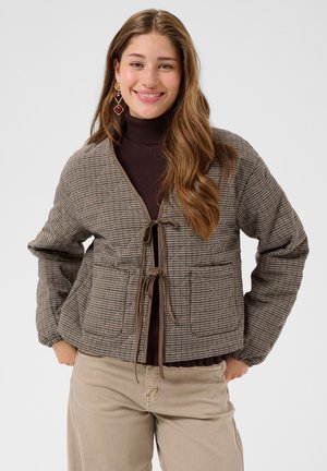 Jeune femme souriante, portant une veste marron à carreaux nouée devant, un col roulé foncé, un pantalon beige et des boucles d'oreilles pendantes colorées.
