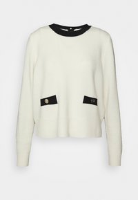 Pull blanc à manches longues avec encolure ronde contrastante noire et rabats de poche noirs, chacun maintenu par un bouton argenté.