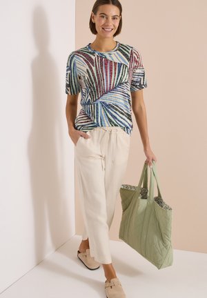 Femme portant un haut multicolore rayé à manches courtes, pantalon large beige, chaussures faciles à enfiler, portant un grand sac cabas vert matelassé.