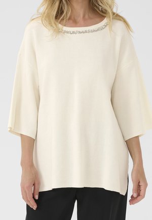 Femme portant un haut en maille ample de couleur crème avec un détail de perles argentées au niveau de l'encolure ronde et des manches larges arrivant aux coudes.