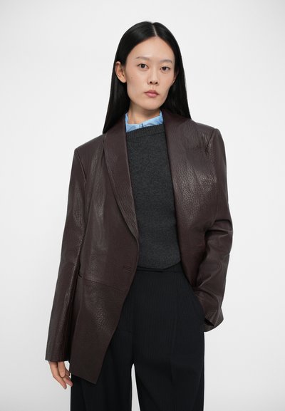 Sacou oversized din piele maro cu un finisaj texturat, asortat cu un pulover gri închis și o cămașă cu guler bleu deschis, plasat pe un fundal simplu.