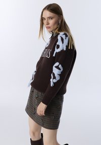 Jonge vrouw met steil blond haar, gekleed in een donkerbruine trui met blauwe patronen en een bruin geruit minirok met versieringen.