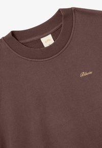 Brauner Sweatshirt mit geripptem Rundhalsausschnitt, weichem Material und kleinem, goldenen, gestickten Logo auf der Brust. Etikett sichtbar im Kragen.
