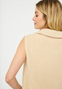 Beige, ärmlös stickad tröja med vikbar krage och ribbad textur, som visar en avslappnad passform och en slät ytdetalj.