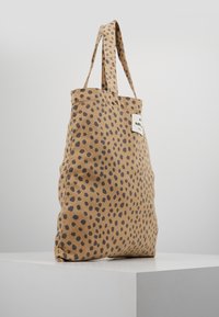 Beige tygväska med svarta oregelbundna prickar och en vit rektangulär etikett med svart text som hänger från korta handtag.