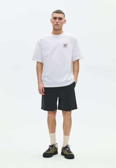 Jeune homme debout, portant une chemise blanche à manches courtes avec un logo noir, un short noir, des chaussettes beiges et des baskets vert-noir.