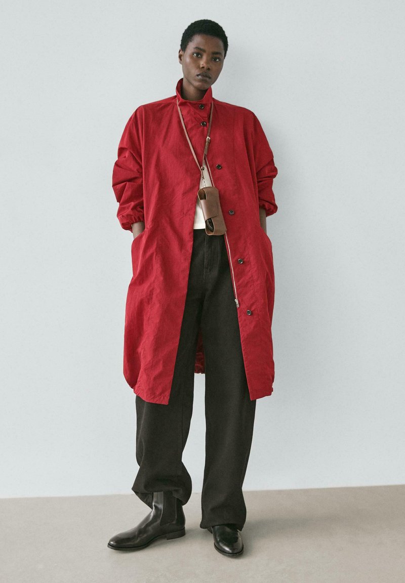 Rote leichte Jacke mit hohem Kragen, Knopfverschluss und Seitentaschen, kombiniert mit dunkel-losen Hosen und schwarzen Stiefeletten.