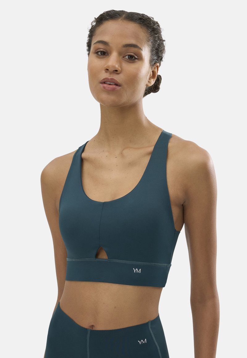 Mujer con el cabello trenzado, vistiendo un top deportivo color azul verdoso y leggings de talle alto, de pie contra un fondo liso y claro.