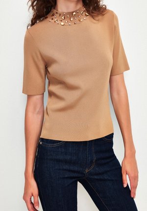Kobieta w beżowej bluzce z krótkim rękawem z ozdobnym, wysokim kołnierzem oraz w ciemnoniebieskich dżinsach, stojąca na jasnym tle.