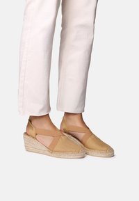 Béžové espadrilky s křížovými elastickými popruhy, tkanou jutovou klínovou podešví a hladkým textilním svrškem. Spárované s lehkými zkrácenými kalhotami.