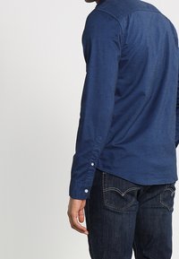 Man i mörkblå långärmad skjorta och mörk denimjeans, stående med ryggen delvis vänd mot kameran mot en neutral bakgrund.