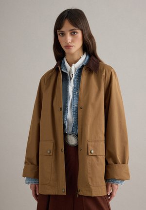 Femme aux cheveux bruns portant une chemise blanche superposée, une veste en jean bleue et un manteau marron surdimensionné avec de grandes poches avant et un col foncé.