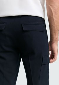 Mann in dunkelblauen Cargo-Hosen mit Klapptaschen auf der Rückseite und an der Seite, kombiniert mit einem in die Hose gesteckten weißen Hemd vor einem schlichten Hintergrund.
