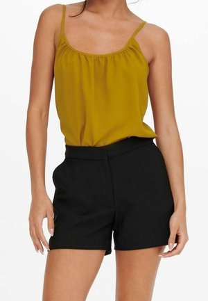 Vrouw draagt een mosterdgeel mouwloos topje met dunne bandjes en zwarte shorts met hoge taille tegen een witte achtergrond.
