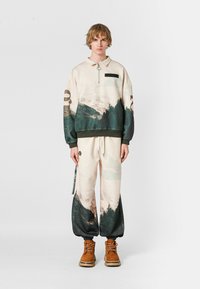 Joven de pie en un suéter de cremallera beige y verde con temática natural y pantalones a juego, con botas marrones, sobre un fondo liso.