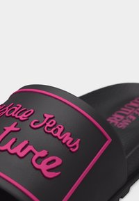 Μαύρο σανδάλι με φάσα, με ανάγλυφο ροζ κείμενο «Versace Jeans Couture» και υφασμένο πέλμα με αντίστοιχη ροζ σήμανση.