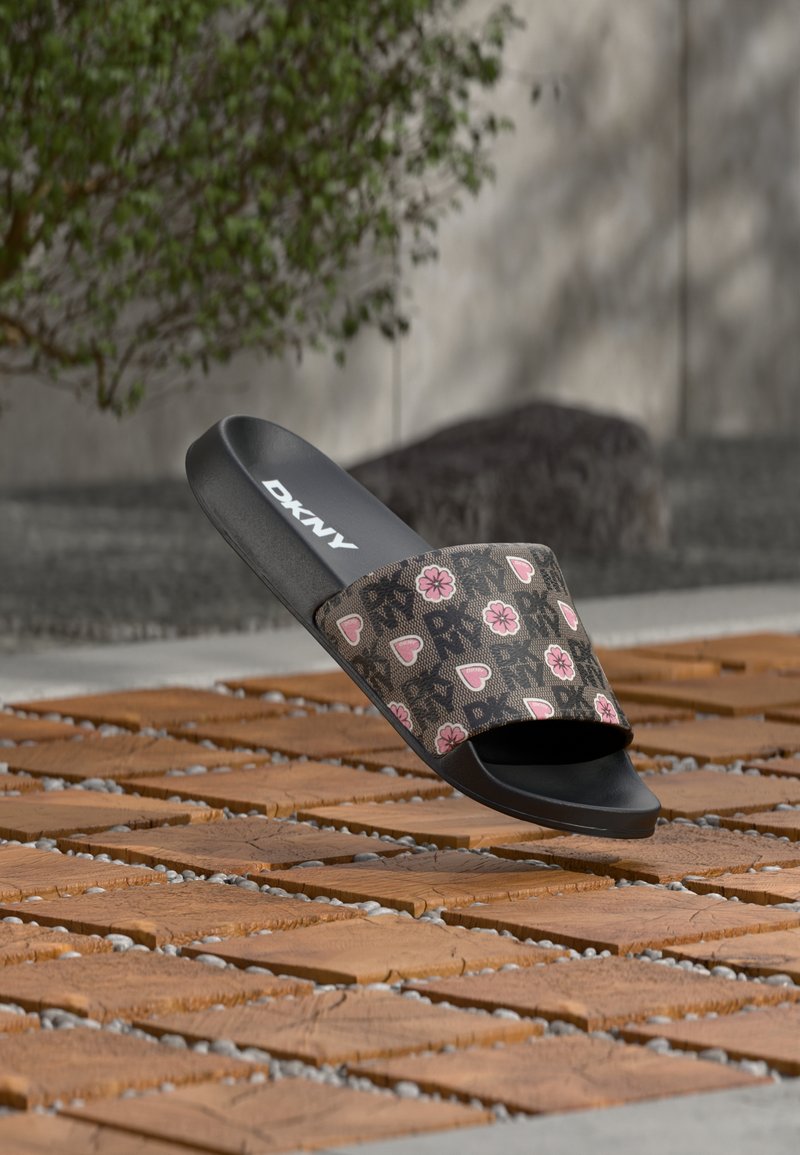 Mule DKNY noire avec une bride marron décorée de cœurs et de fleurs roses, posée sur des carreaux en bois en extérieur.