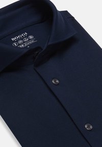 Polo majica mornarsko modre barve z teksturirano tkanino, z usnjeno ovratnikom in tremi gumbi. Ima tudi oznako blagovne znamke znotraj ovratnika.