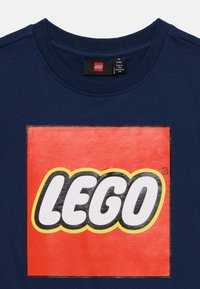 T-shirt en coton bleu marine avec un grand logo rectangulaire rouge portant le mot "LEGO" en typographie grasse blanche et noire. Encolure ronde.