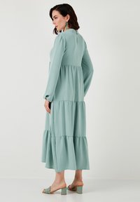 Flowy, tiered maxi dress in soft mint green fabric. Features long sleeves, a round neckline, and a back button closure. Flat sandals with block heels.