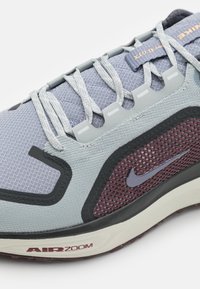La basket Nike présente une tige en mesh gris, des accents bordeaux, des éléments réfléchissants et une semelle extérieure en caoutchouc noir texturée avec le logo « AIR ZOOM ».