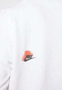 Vit sweatshirts med en svart och orange Nike-logotyp med en gradienteffekt, texturerat material och en klassisk rundhalsdesign.