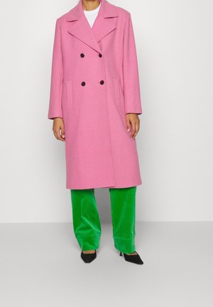 Manteau double boutonnage en mélange de laine rose avec grands revers, boutons noirs, deux poches latérales ; associé à un pantalon en velours vert et des talons noirs.