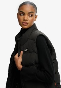 Chaleco acolchado negro con cuello alto, cierre de cremallera y parche con logo. Textura suave, diseño ligero y silueta ajustada.