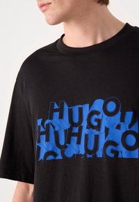 Camiseta de algodón negra con texto azul en negrita en un estilo en capas, 3D que dice "HUGO", de manga corta y con diseño de cuello redondo.