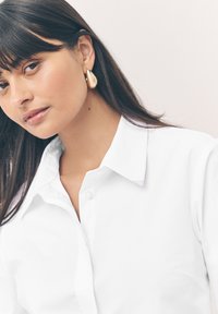 Chemise blanche boutonnée avec un col classique, une texture lisse et une brillance subtile. Des boucles d'oreilles en forme de goutte en or complètent le look.